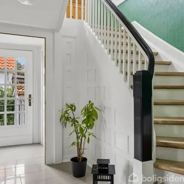 En elegant entré med trappe, grøn væg, og hvidt panel. En plante i sort potte og to lanterner står ved trappen. Dør med glasruder fører ud til solrig gårdhave.