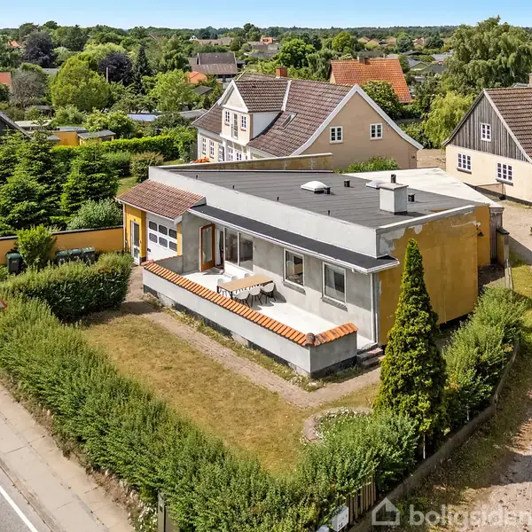 Moderne hus med fladt tag ligger langs en vej, omgivet af frodige buske og træer. Huse i traditionel stil ses i baggrunden, hvilket skaber en hyggelig nabolagsstemning.