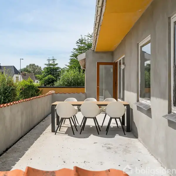 En terrasse med et træbord og seks hvide stole står omkranset af en mur med orange tagsten. Døre fører ind i huset, og omgivelserne består af grønne buske og huse.