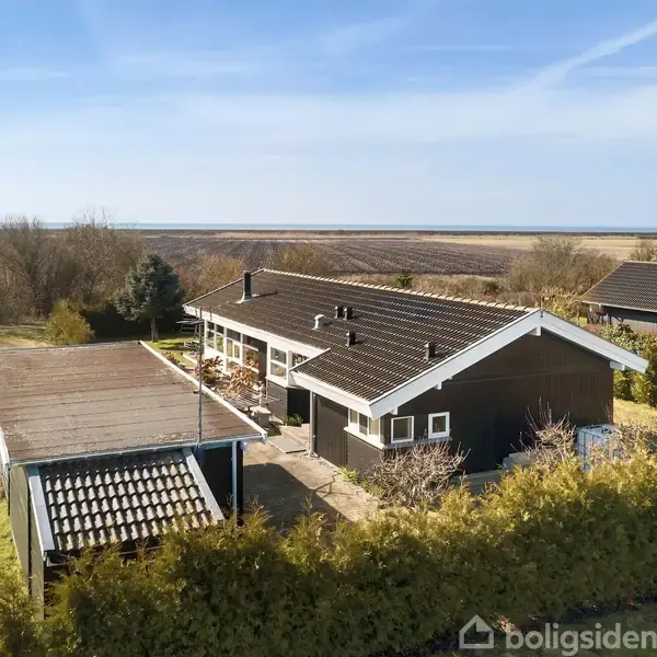 En mørk træbeklædt villa står stille i et åbent, frodigt landskab med udsigt til marker og hav. Huset har et tilhørende carport og er omgivet af grøn beplantning.