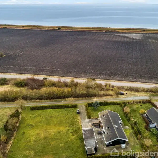Huse ligger stille på græsplæner, omgivet af træer, foran en stor, pløjet mark. Havet strækker sig i horisonten under en skyet himmel.