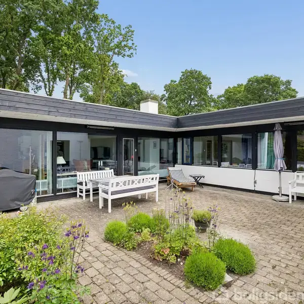 En gårdhave med flisebelægning, omkranset af et L-formet moderne hus med store vinduer. Bænke og en grill er placeret ved husets vægge, omgivet af grønne planter.