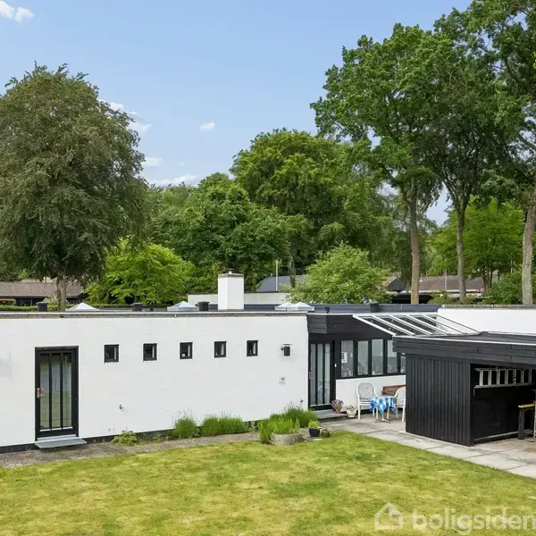 Et moderne, hvidt hus med fladt tag står i en have omkranset af høje træer. Der er en terrasse med glasoverdækning og udemøbler. Ingen synlig tekst i billedet.