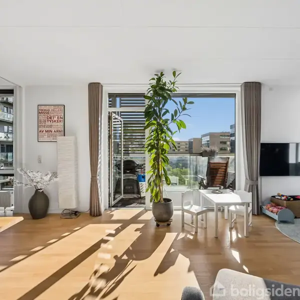En høj plante står i en lys stue med en træbord og stole. Et TV hænger på væggen ved siden af legetøj og en balkon er synlig udenfor.