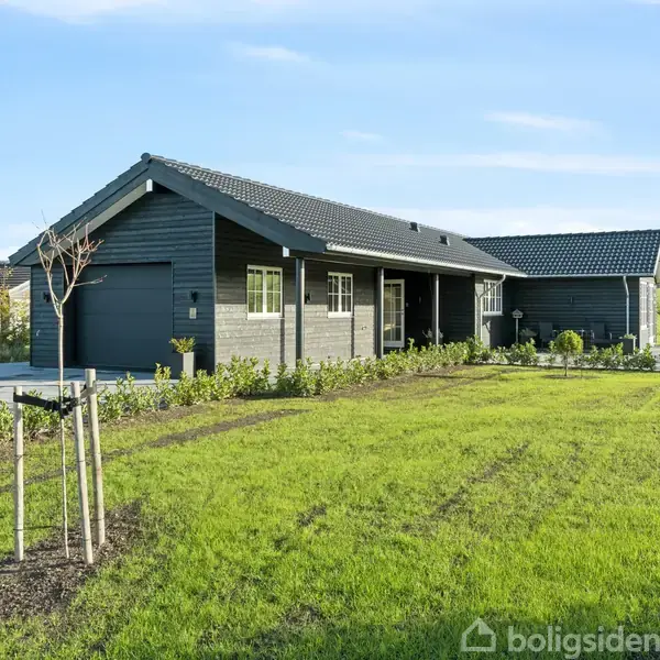 Et moderne, sort træhus står stille på en velplejet grøn græsplæne med unge træer. Huset har en tilhørende garage og er omkranset af en veltrimmet hæk, beliggende i et landområde.