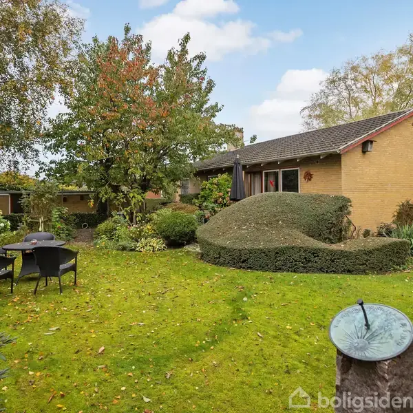 Et gult murstenshus står stille i en frodig have med klippede buske og træer. Foran huset er der en græsplæne med sorte havemøbler og et solur i forgrunden.