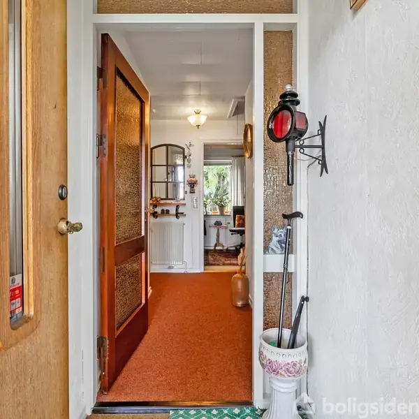 Dør åbner ud til en entré med rødt gulvtæppe. En hat hylder med paraplystativ står til højre, og malerier hænger på væggen. En stue anes i baggrunden.