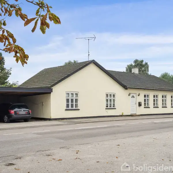 Et lysegult én-etagers hus med gråtag holder til i et byområde. Ved siden af huset er der en carport med en parkeret bil. Træer og andre bygninger er i baggrunden.