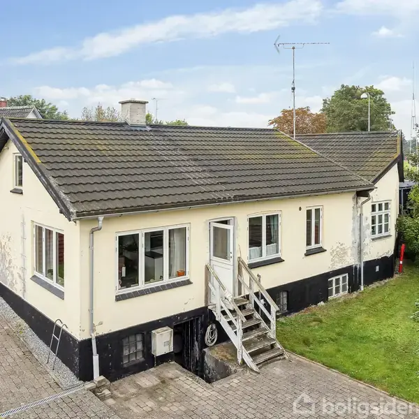 Et gulmalet hus med sort sokkel ses med trappe til hoveddør. Det er omgivet af en gårdhave med fliser og en græsplæne. Nabohuse ses i baggrunden.