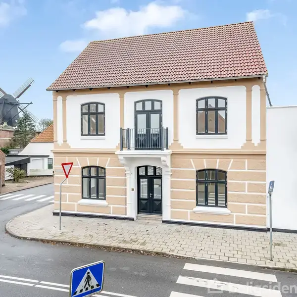 Et stort, hvidt to-etagers hus med rødt tag står ved en vej, hvor der er et fodgængerfelt og et skilt med "Vigepligt." I baggrunden ses en mølle.