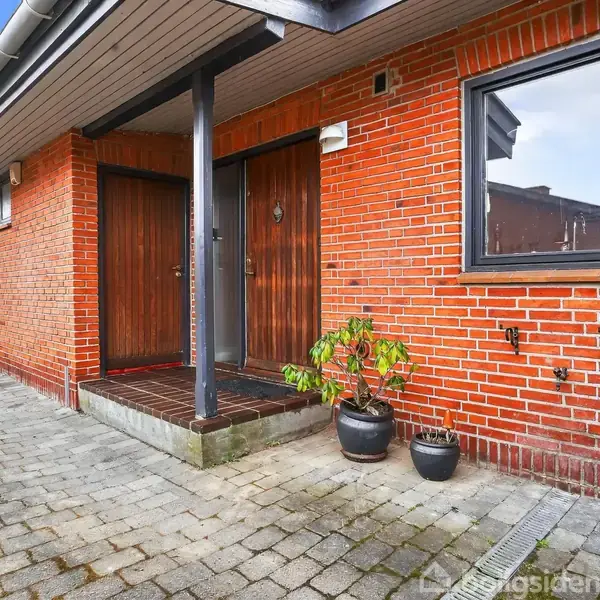 Rødt murstenshus med to trædøre er hovedfokus. To sorte potter med planter står foran dørene. En indkørsel med brosten omgiver huset. Ingen tekst er synlig på bygningen.