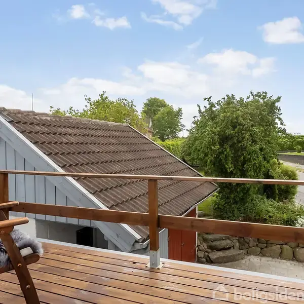 Stol med pels sæde står stille på en træterrasse med udsigt til en lille grå bygning, træer og en vej under blå himmel med skyer.