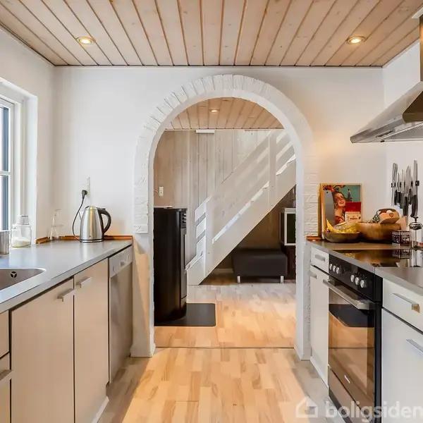 Et moderne køkken med grå skabe. Øelementer inkluderer en vask, komfur og emhætte. Et buet indgangsparti fører til en stue med trægulv og gulvtæppe. Lysindfald fra et vindue.