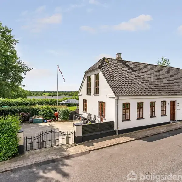 Hvidt hus med sort tag står ved gade med flagstang i forhaven. Omkringliggende buske og træer skaber en grøn baggrund med blå himmel.