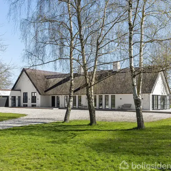 Huset har stråtækt tag og hvide vægge, står stille omgivet af græsplæner og løvtræer. Grussti leder til garager. Blå himmel skaber en rolig atmosfære.