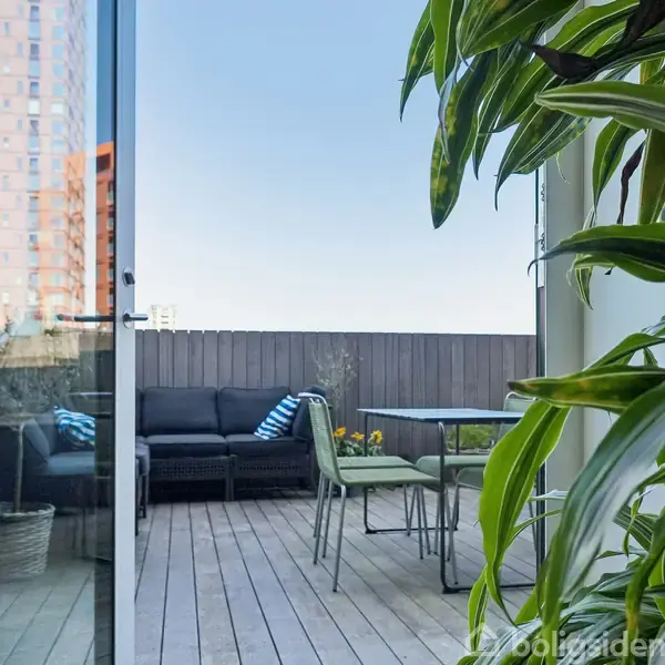 En terrasse med trædæk har et bord og stole. I baggrunden står en sort sofa med stribede puder. Planter omgiver området, og en høj bygning ses i baggrunden.