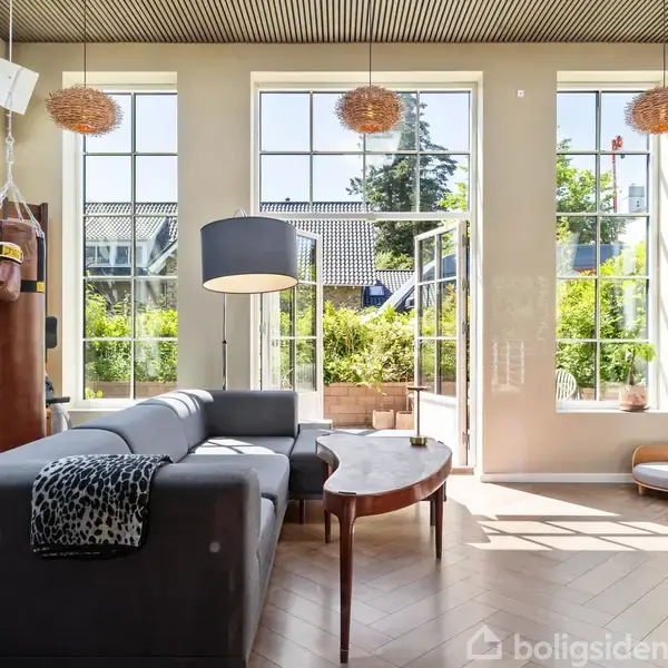 Sofaen står i stuen ved store vinduer, hvor sollys strømmer ind. En boksepude hænger fra loftet, og dørene åbner ud til en frodig have.