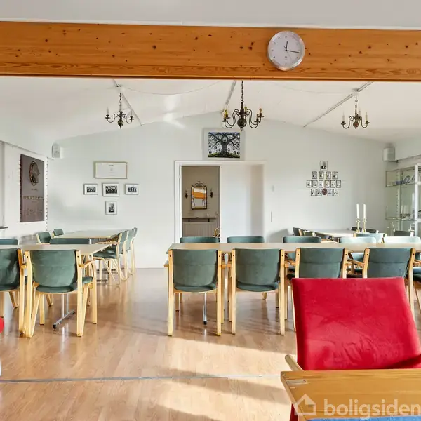 En spisestue med opstillet borde og stole, hvor borde danner en U-form. Rummet er lyst, med en stor vindue og lysekroner i loftet.