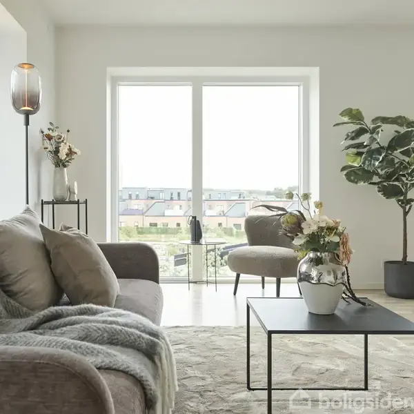 Sofa med tæppe står i en stue, omgivet af planter og et sofabord med blomster. Store vinduer giver udsigt til byen. Lampen står tændt ved siden af en vase.