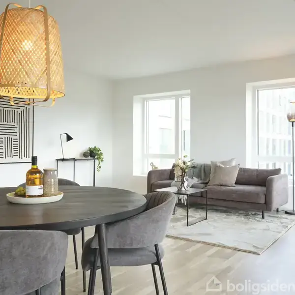 Spisebord med fire stole står i stue. Rund lampe hænger over bordet. Sofa med puder står ved vindue. Væglampe og plante i hjørnet. Maleri på væggen.