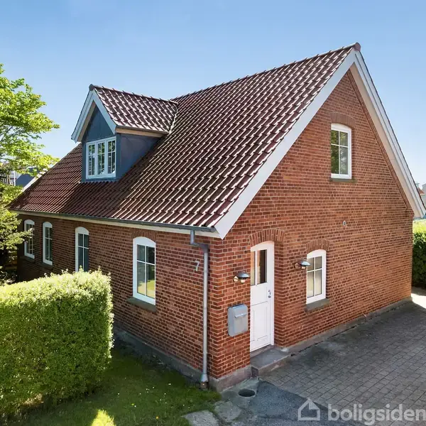 Rødt murstenshus med skråt tag og kvist, står stille i solskin. Omgivet af grøn hæk og belagt indkørsel, med udsigt til marker og en fjern horisont.