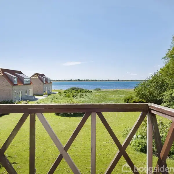 En træbalkon med udsigt over en grøn have leder blikket mod et blåt hav og klare himmel. Til venstre ses murstenshuse med røde tage.