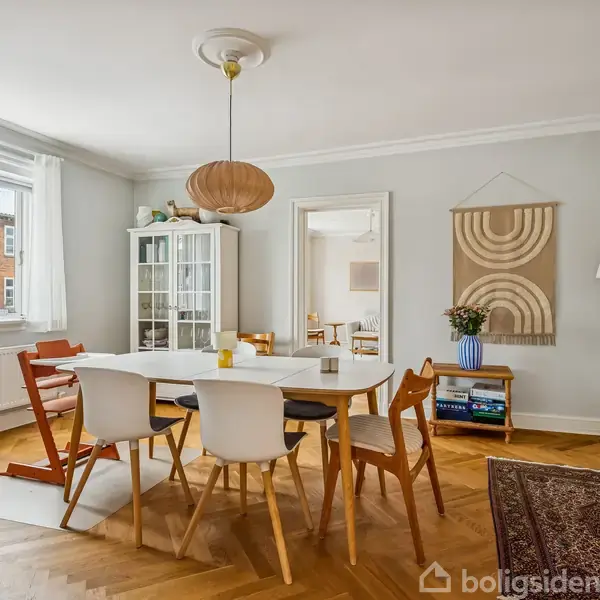 En spisestue med et rektangulært bord omgivet af forskellige stole. En lampe hænger over bordet. Rummet har lyse vægge, et vindue med gardiner og dekorativt tæppe på trægulvet.