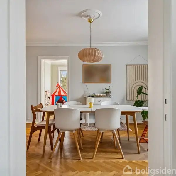 En spisebordsgruppe med hvide stole står i et lyst rum med parketgulv. En lampe hænger over bordet. I baggrunden ses et farverigt legetelt og planter.