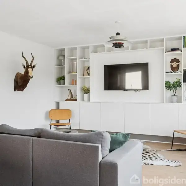 Gråt sofaarrangement vender mod hvid skænk med indbygget TV. Hylder viser dekorationer. Væggene prydes af kunst og hjortetrofæ. Rummet har træstole og et mønstret tæppe på gulvet.
