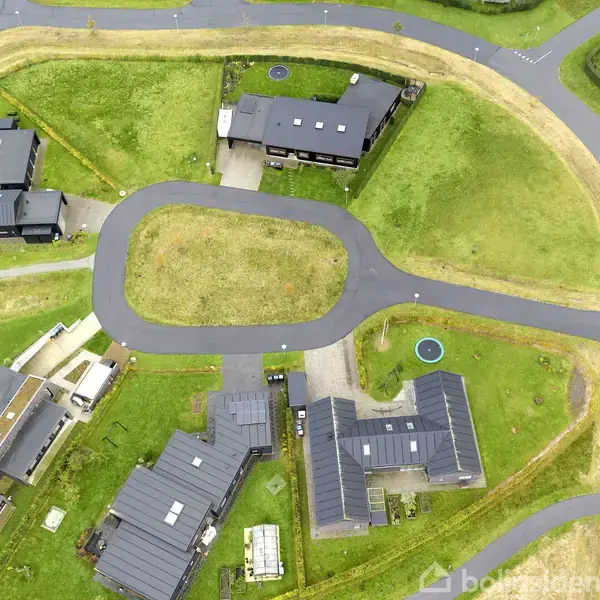 Moderne huse med flade tage ligger spredt rundt om en oval vej, omgivet af grønne græsarealer og mindre stier, i et beboelsesområde med klar struktur.