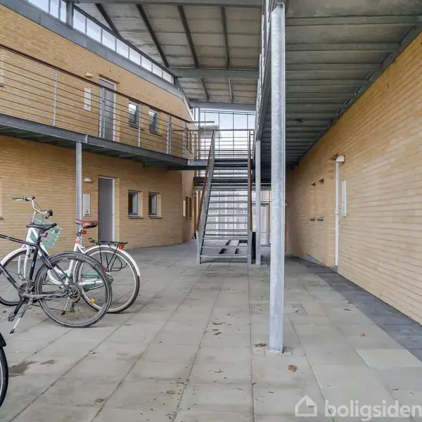 Cykler står parkeret i en gårdhave med flisebelægning. Omgivet af en moderne bygning med trapper. Adresseplade viser: "6".
