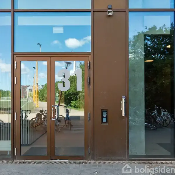 Indgangsdør med vinduer viser tallet "31". Udenfor står cykler, og spejlingen i glasset viser en park med træer under en blå himmel med skyer.