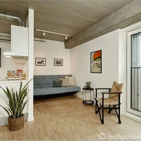 Stue med sofa og stol, hvor sofaen er placeret mod en hvid væg med kunst og plakater. Store vinduer til højre slipper lys ind. Planter dekorerer rummet.