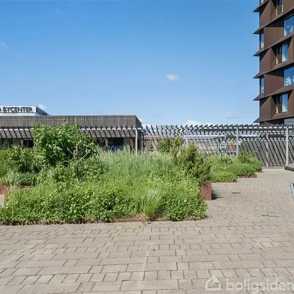 En gårdhave med grønne planter og bænke er omgivet af murede bygninger. Skiltet viser "Næstved Bycenter" i baggrunden under en blå himmel.