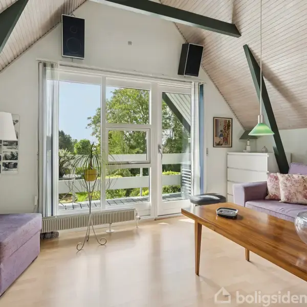 Stue med lilla sofa og sofabord med skål, set mod store vinduer ud til have. Hvidt skab, grøn lampe og indrammede billeder på væggene tilføjer dekoration.