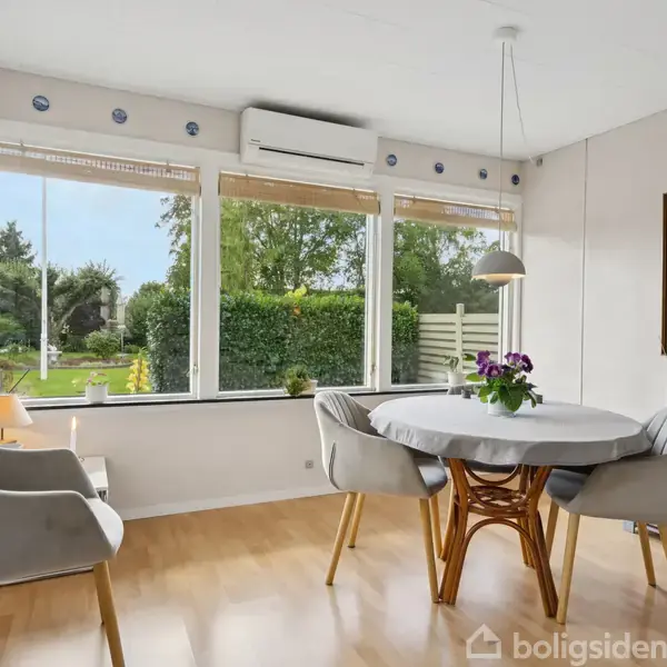 Et rundt spisebord står med fire stole i en lys stue, der har store vinduer med udsigt til en grøn have. En lampe hænger over bordet. En plante og et maleri pynter.