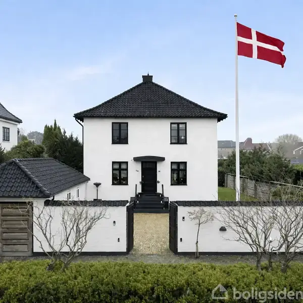 En hvid villa står foran en dansk flagstang i en velplejet have. Huset har et mørkt tag og symmetrisk facade, omgivet af en lav mur med grønt buskads.