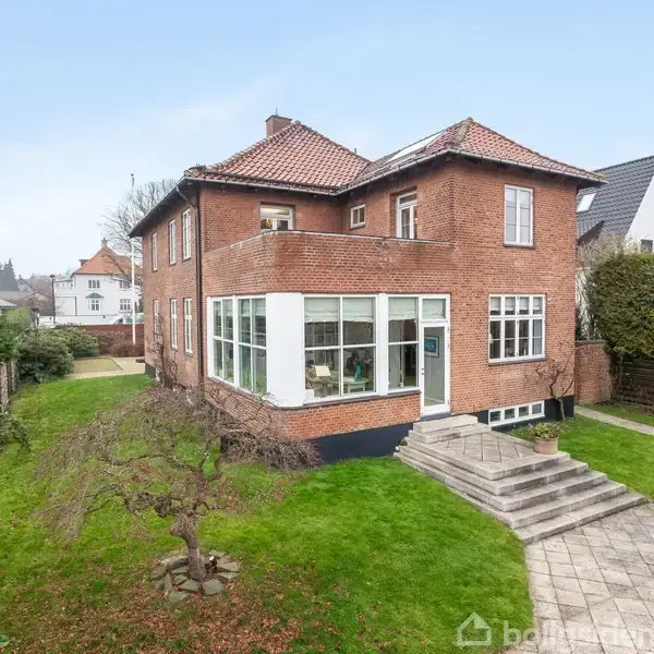 Rød murstensvilla med rødt tegltag, står stille på en velplejet græsplæne. Omkranset af hække og flisebelagte stier i en forstad. Vinduerne er store og indbydende.