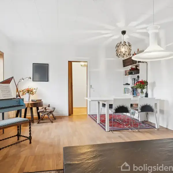 Et lyst stueinteriør med et klaver ved siden af et vindue. Et spisebord står nært en bogreol med blomster. Rummets atmosfære er hyggelig og indbydende.