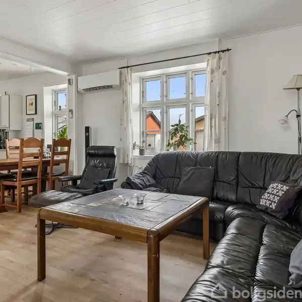 En sort lædersofa står rundt om et sofabord i en lys stue med trælaminatgulv. Et spisebord i træ står til venstre ved siden af et åbent køkken.