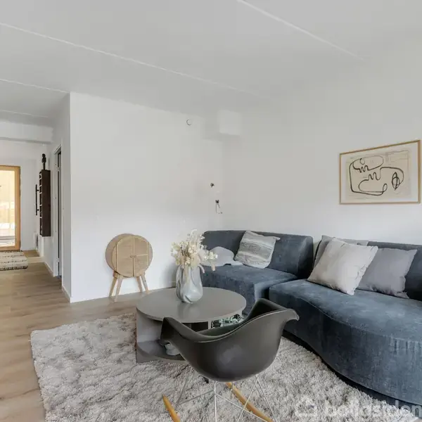 Gråt sofaarrangement står stille i en lys stue med trægulv. Et lille rundt bord med vase står foran sofaen. Køkken til venstre, maleri på væggen.