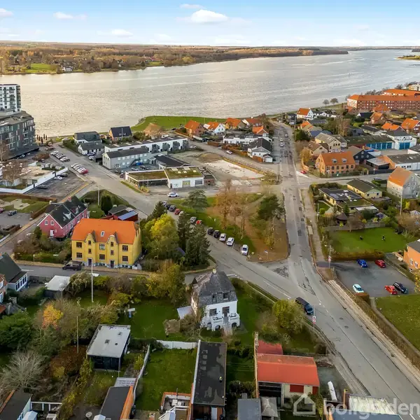 Luftfoto af en by ved vandet med huse i forskellige farver, en vej der løber gennem kvarteret, og lejlighedsbygninger i baggrunden ved floden.