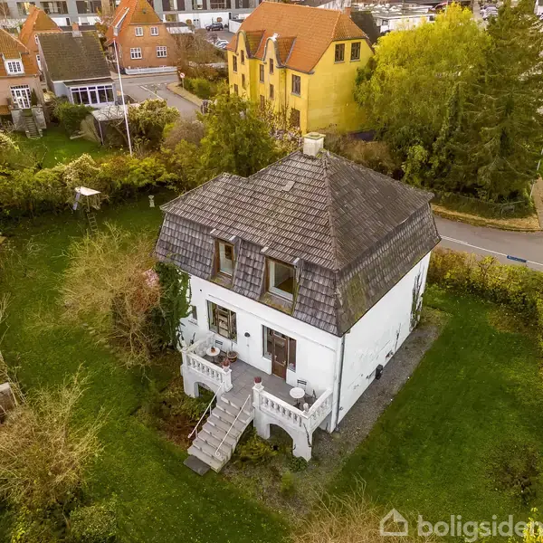 En firkantet hvid villa med gråt tag står omgivet af en frodig have. Villaen har to store vinduer på tagetagen og en trappe ved hovedindgangen. I baggrunden er der flere farverige huse og en gade.