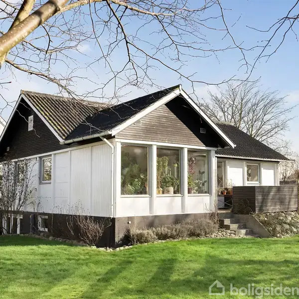 Et hvidt hus med mørkt tag står i en have med grønne plæner og træer. Store vinduer ses på forsiden, og en træterrasse ligger til højre.