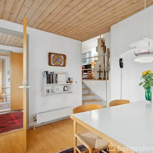 Et spisebord med hvide overflader og træstole står i et lyst rum. En moderne lampe hænger over bordet. Væggen har en hylde med bøger, og en grøn vase med blomster.