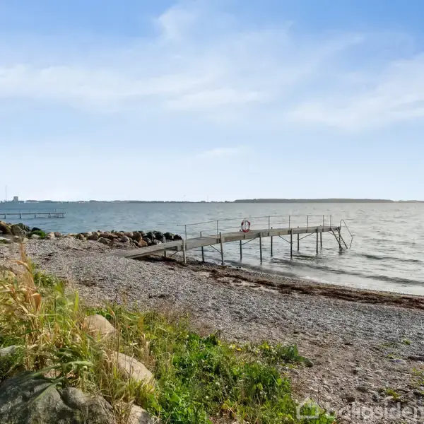 En træbro strækker sig ud i vandet fra en stenet strand med græs og klipper, under en blå himmel med lette skyer.