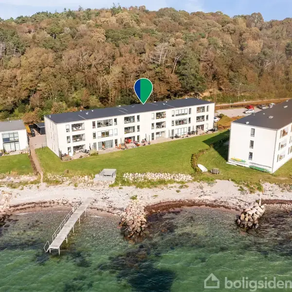 Bygning står ved vandet med en fantastisk udsigt, markeret af en blå nål. Græsarealer og en mole omgiver bygningen. Skov danner baggrunden.