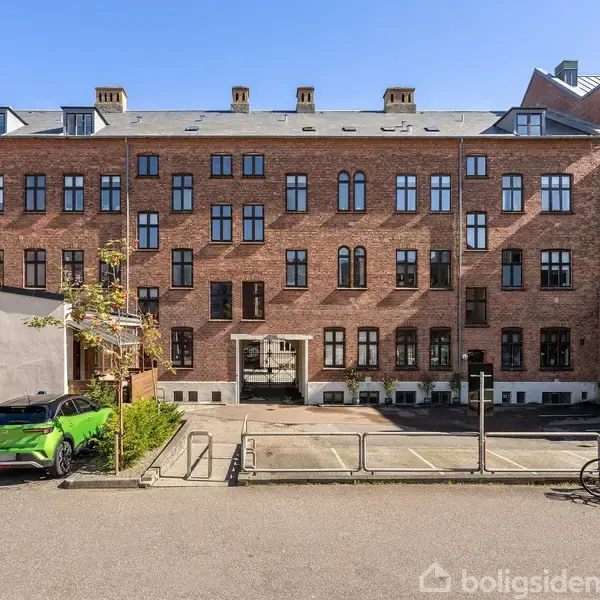 En rød murstensbygning med vinduer står stille i sollys. En grøn bil er parkeret foran bygningen. Cykler er opstillet ved siden af nogle parkeringspladser.