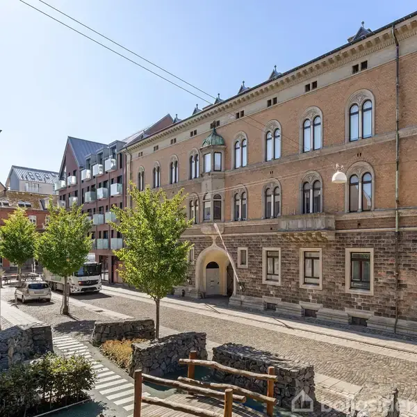 En historisk bygning med buede vinduer står stille på en brostensbelagt gade. Omkring bygningen er moderne lejlighedskomplekser og parkerede biler. Træer er placeret langs vejen.
