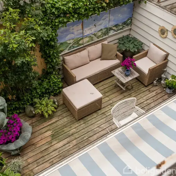 Terrasse med lounge-sofa og bord i flet, omgivet af planter og blomster. En skulptur i hjørnet sidder ved en blomsterbeplantning, mens væggene er dekoreret med kunst og solspejle.